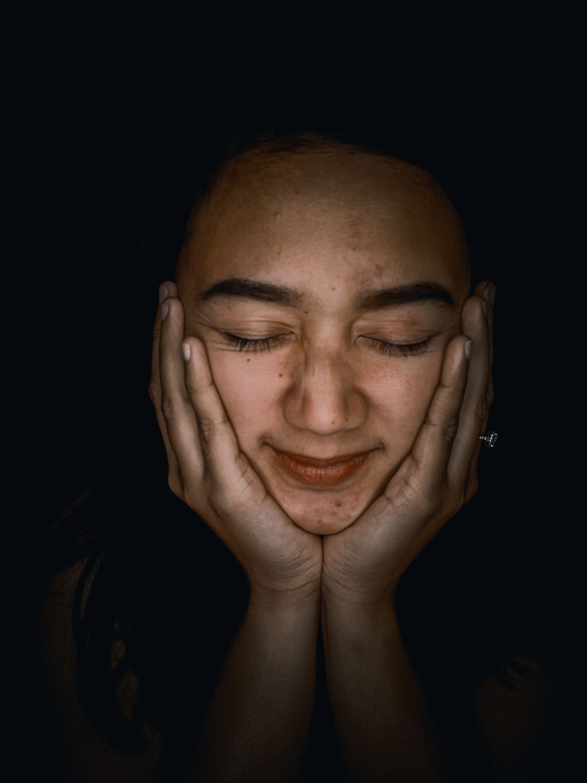A woman with her eyes closed, hands resting on her face, lit softly against a dark background